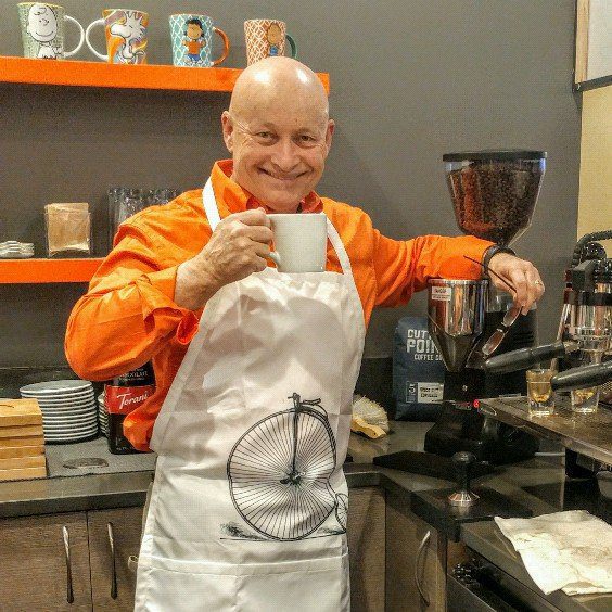 A man in an orange apron holding a cup of coffee while showcasing e-bikes accessories.