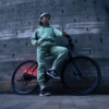 A person in a green tracksuit and helmet stands with a Soltera 2.5 Ebike, featuring a red light, against a concrete wall.