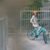 A person in a coral helmet and gray tracksuit stands with a Soltera 2.5 Ebike on a pedestrian bridge, looking back at the camera.