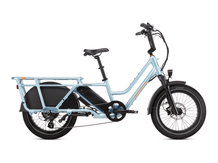 A light blue, electric cargo bike with a step-through frame, black saddle, and integrated rear cargo rack. The bike has front and rear fenders and an electric motor mounted in the rear wheel hub.