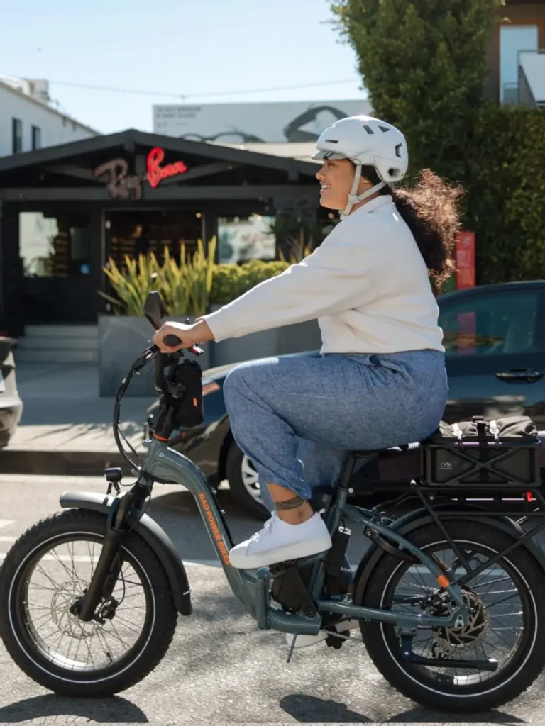 A person wearing a white helmet and casual clothing rides an electric bike on a sunny day in an urban area, with buildings and plants in the background.