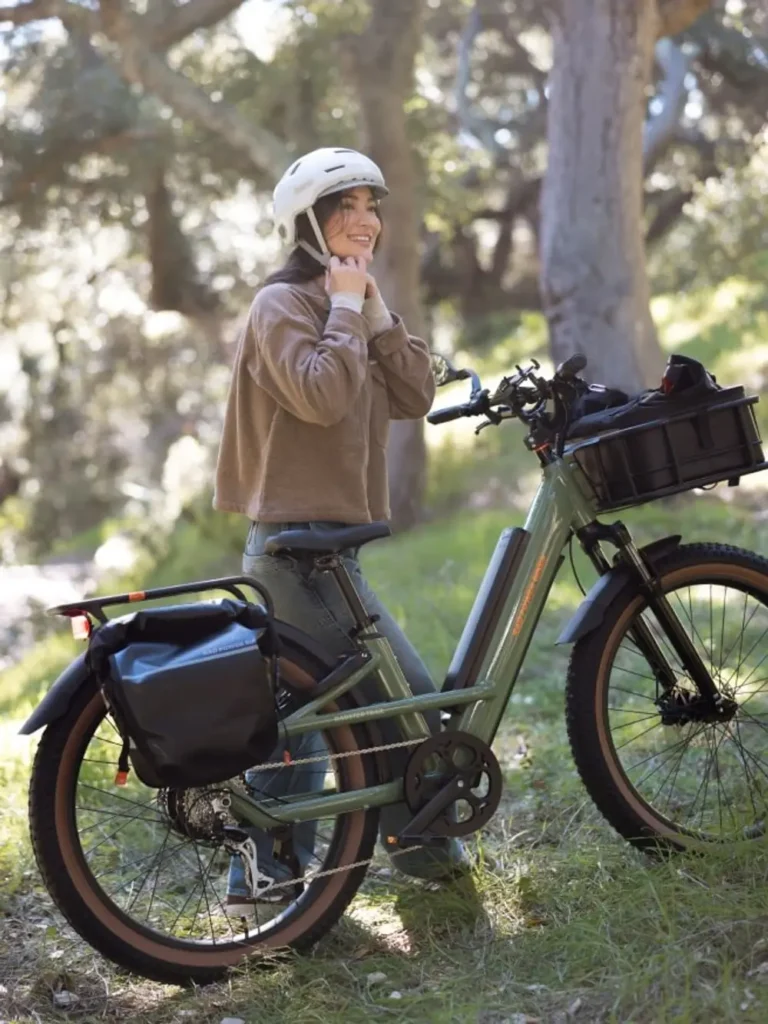 A person wearing a helmet adjusts its strap while standing beside an e-bike on a forest trail.