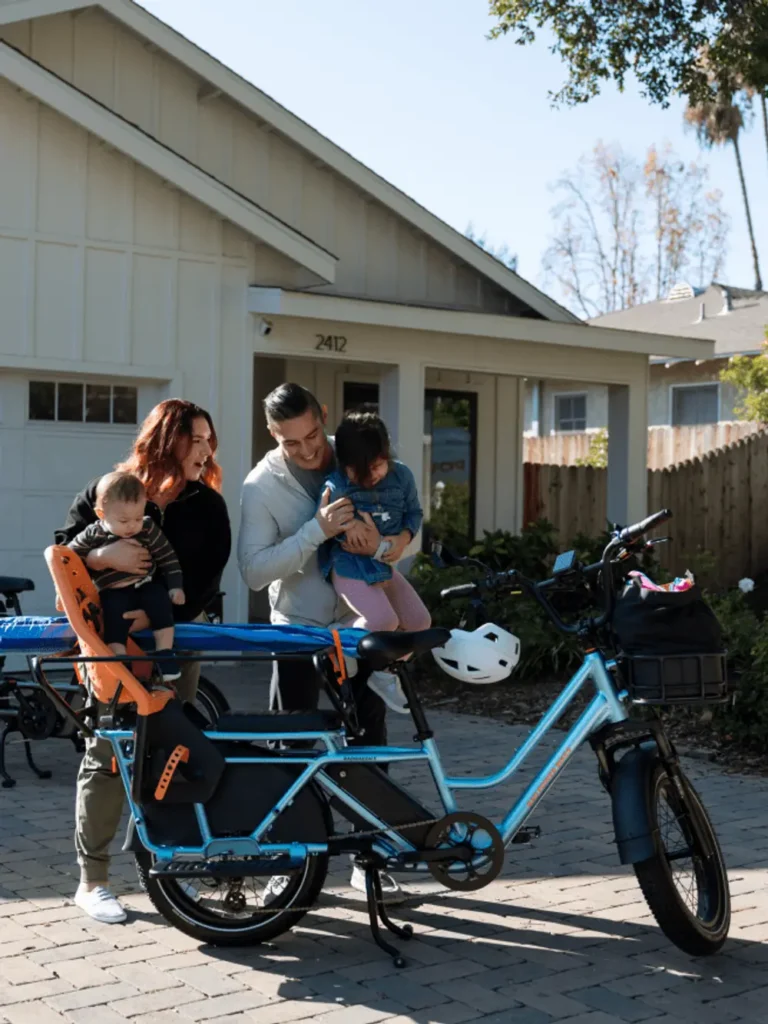 A family stands in front of a house with two adults holding their young children next to a blue e-bike equipped with child seats.