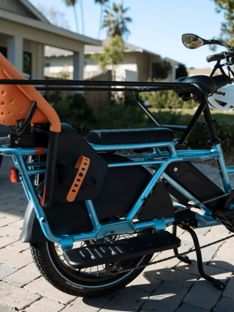 Close-up of a blue cargo bike with an orange child seat mounted on the back. The bike is parked on a paved surface with a house and trees visible in the background.