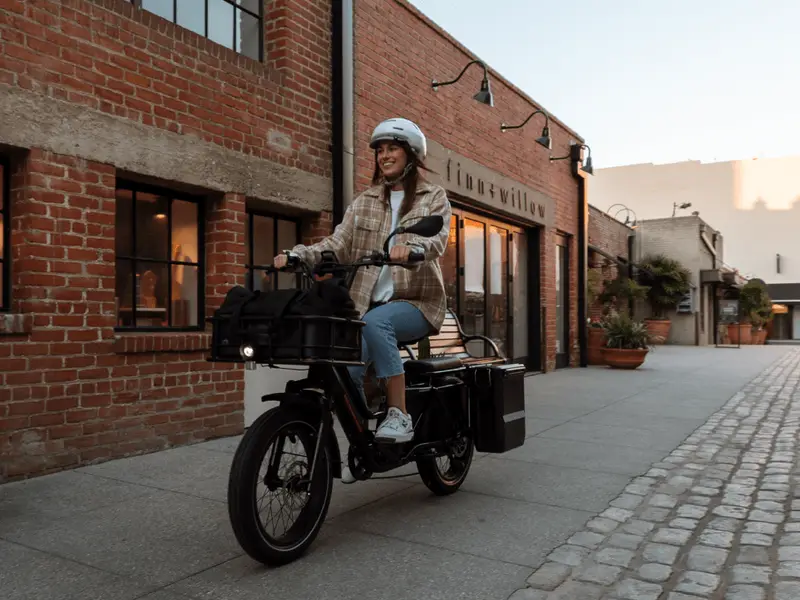 A person rides an electric cargo bike on a cobblestone street, wearing a helmet and casual clothing, with brick buildings in the background.