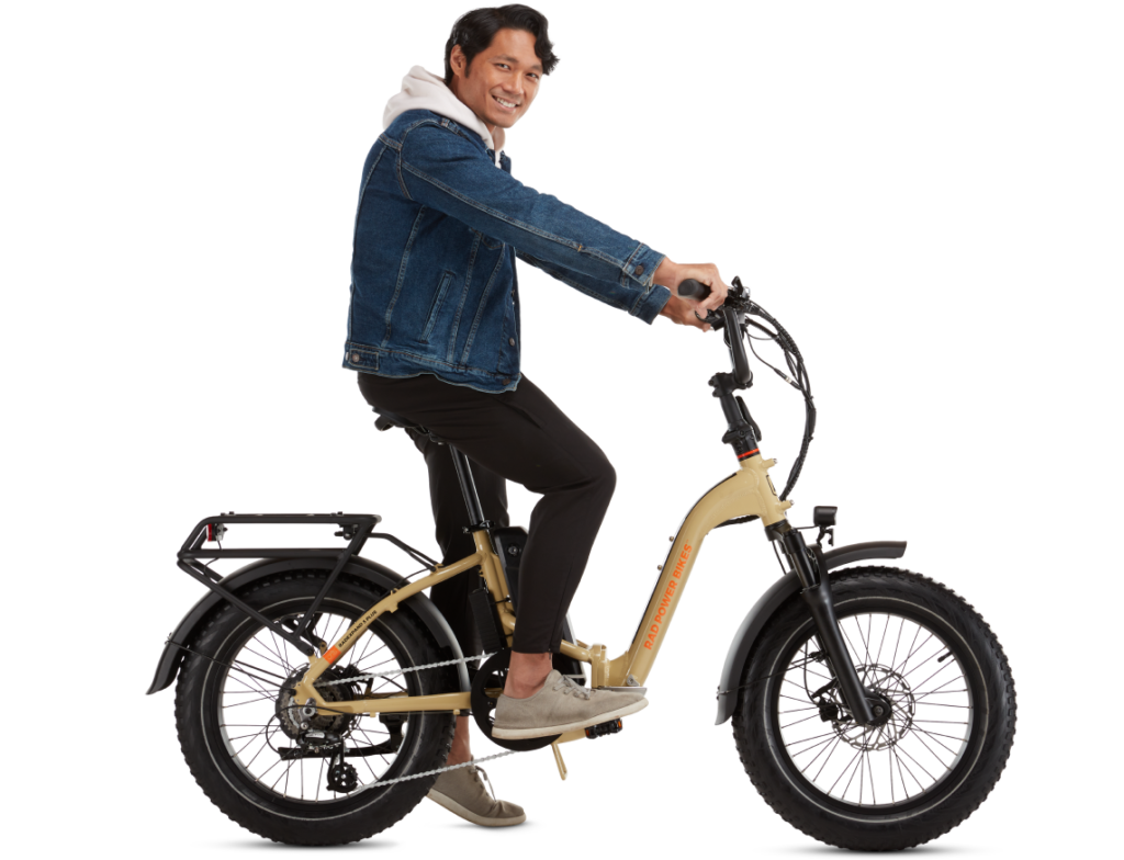 A man smiling rides a beige electric bike while wearing a denim jacket, hoodie, black pants, and gray shoes.