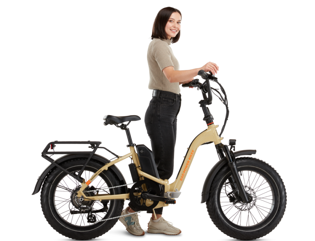 A woman stands next to a beige electric bike with her hands on the handlebars. She is smiling and dressed in a short-sleeve shirt and jeans. The bike features thick tires and a battery pack.