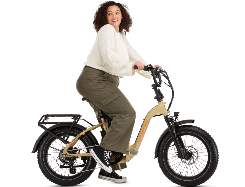 A woman wearing a white sweater and olive green pants rides a beige e-bike against a white background.