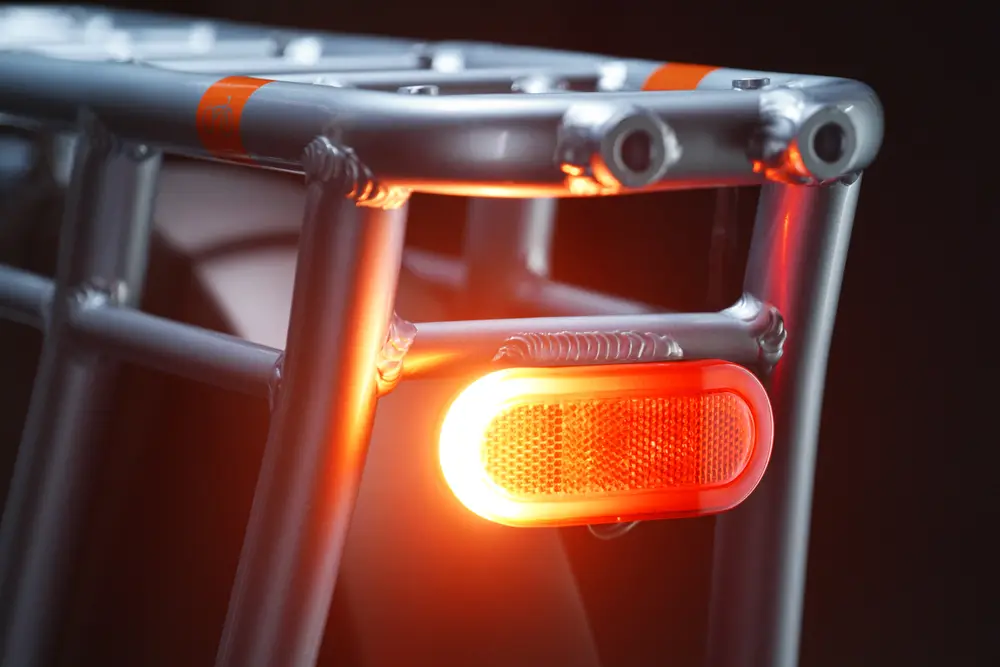 Close-up of a bicycle rack with a lit red reflector at the rear against a dark background. The metal framework is visible, showing its sturdy construction.