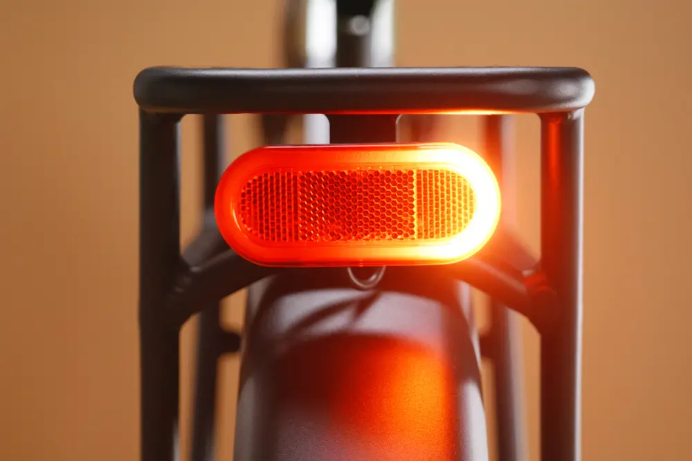 Close-up of a bicycle's rear red reflector light, attached to the bike's frame, against a neutral background.