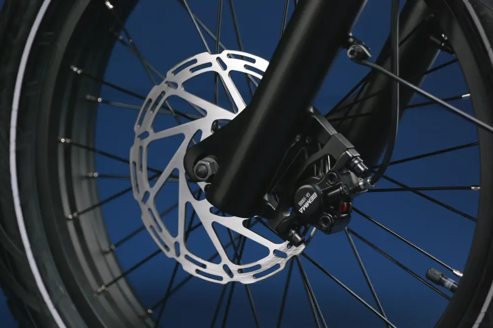 Close-up view of a bicycle's disc brake system mounted on the front wheel, featuring a metallic rotor and caliper against a blue background.