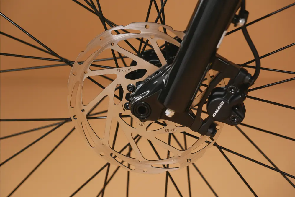 Close-up of a bicycle's front wheel showing a Tektro brake rotor and caliper. The disc rotor is silver with multiple holes, and the surrounding wheel spokes are black. The background is a beige color.