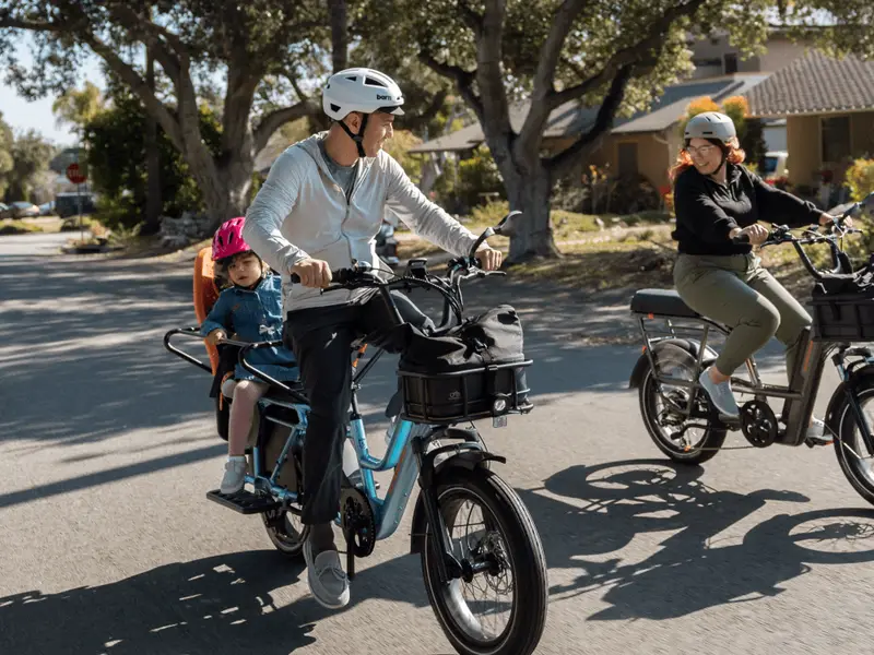 A man rides an electric cargo bike with a child in a rear seat. Another person rides a smaller electric bike alongside on a sunny residential street. Both wear helmets.