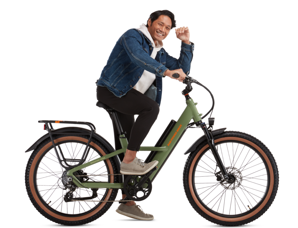 A person sits smiling on a green electric bike, wearing a denim jacket, white hoodie, dark pants, and gray shoes against a plain white background.