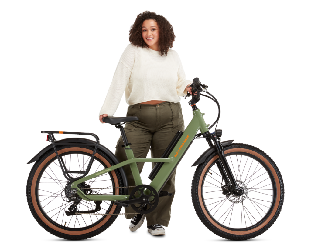 A person with curly hair, wearing a white sweater and green pants, stands holding a green electric bike with brown tires and a rear cargo rack.