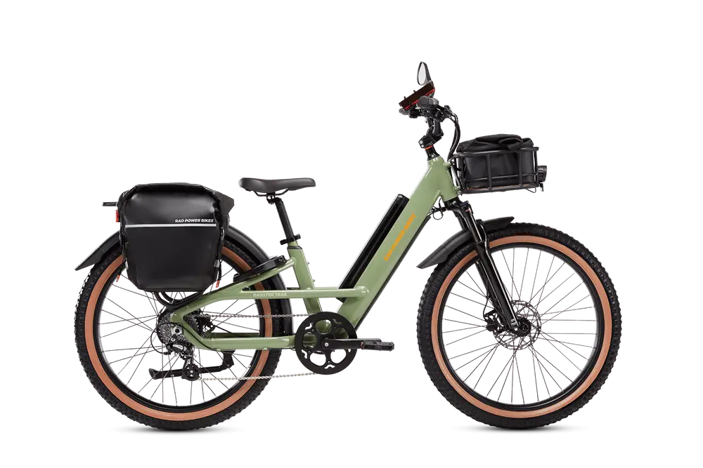 A green electric bicycle with black saddlebags on the rear and a black basket on the front. The bike has wide tires, disc brakes, and a step-through frame.