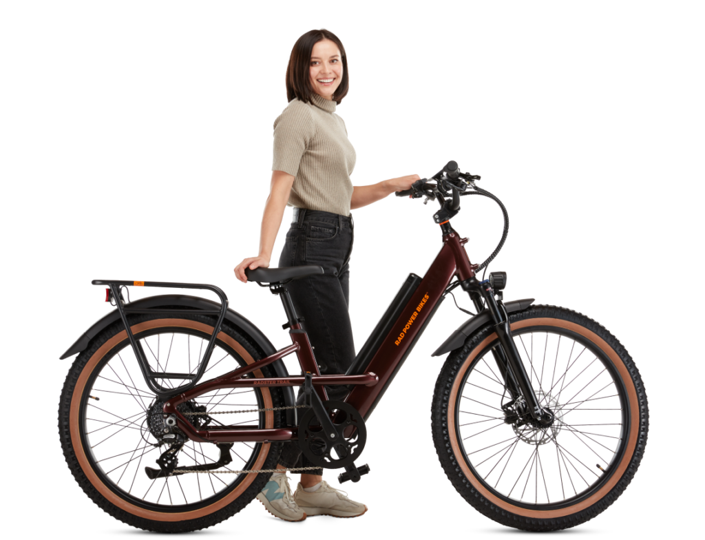 A woman stands next to a maroon electric bicycle, smiling at the camera. The bike features wide tires, a mounted battery, and a rear cargo rack.