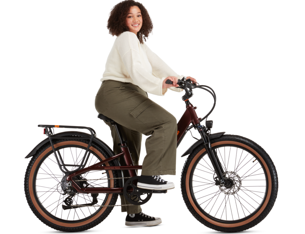 Person with curly hair riding a brown bicycle, wearing a white sweater, olive green pants, and black sneakers.