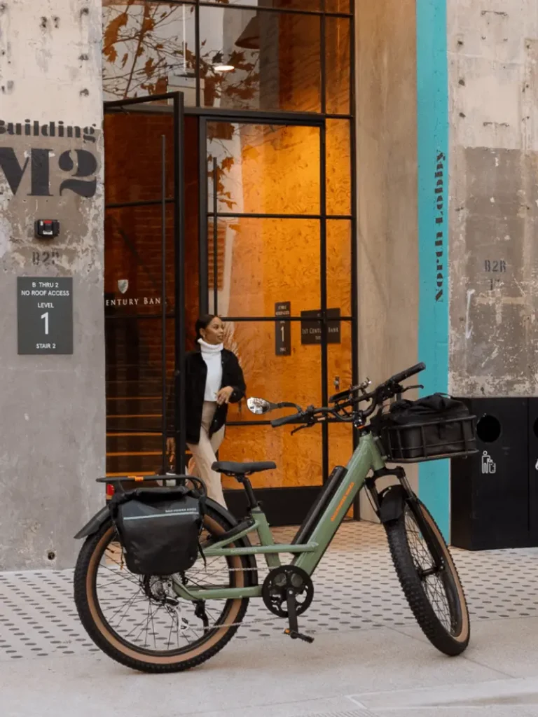 A woman exits a glass door of a building labeled "M2" and "North Lobby." A green electric bike with panniers is parked on the sidewalk in front.