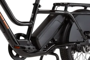 Close-up view of an electric bicycle's battery mounted on the frame, with parts of the rear wheel and cargo rack visible. The bike frame has a "RADWAGON4" label on it.