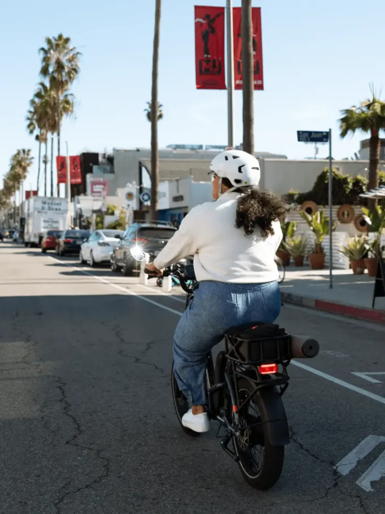 A person wearing a white helmet and white sweater rides a motorcycle on a city street with palm trees and parked cars on either side.