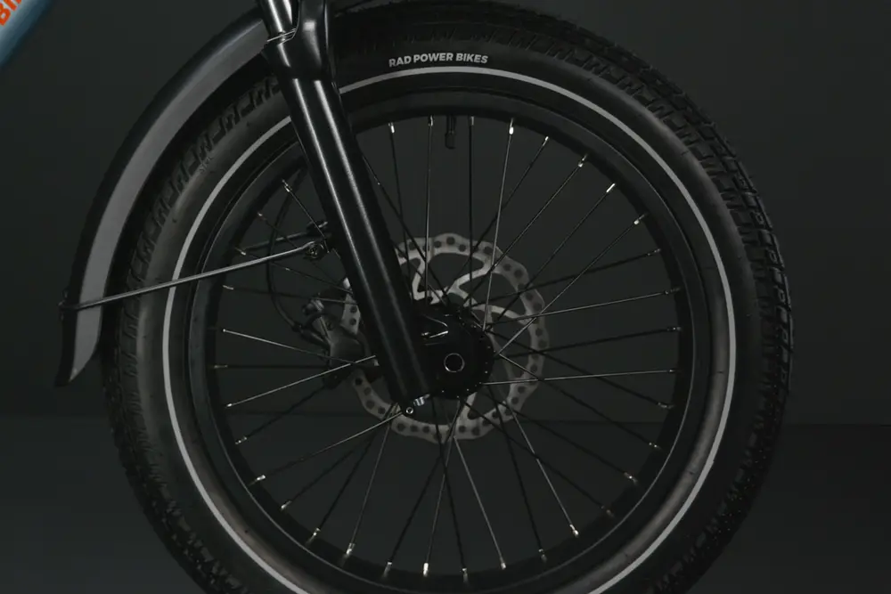 A close-up of a bicycle's front wheel with disc brakes, black spokes, and the brand "Rad Power Bikes" visible on the tire.