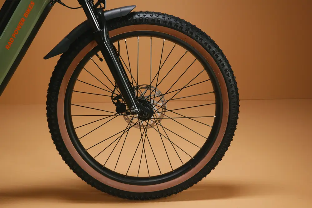 Close-up of a bicycle front wheel with knobby tires and disc brakes in front of a tan background.