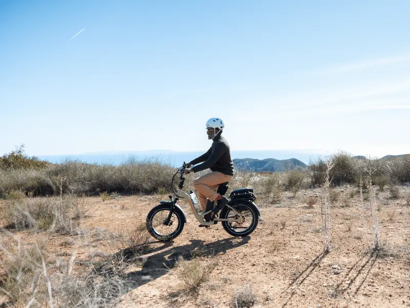 A person wearing a helmet rides an electric bike on a dirt trail in a dry, open landscape with hills and clear skies in the background.