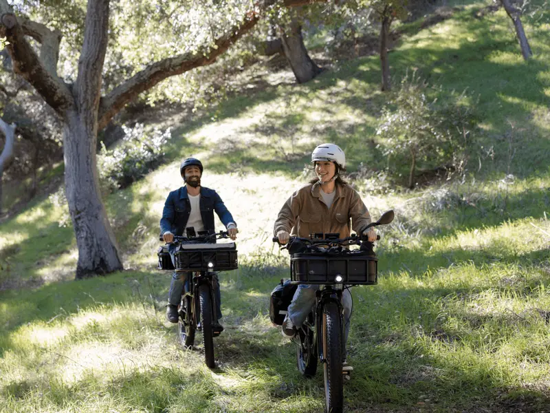 Two people ride electric bicycles through a forested area with green grass and trees, both wearing helmets and casual clothing.