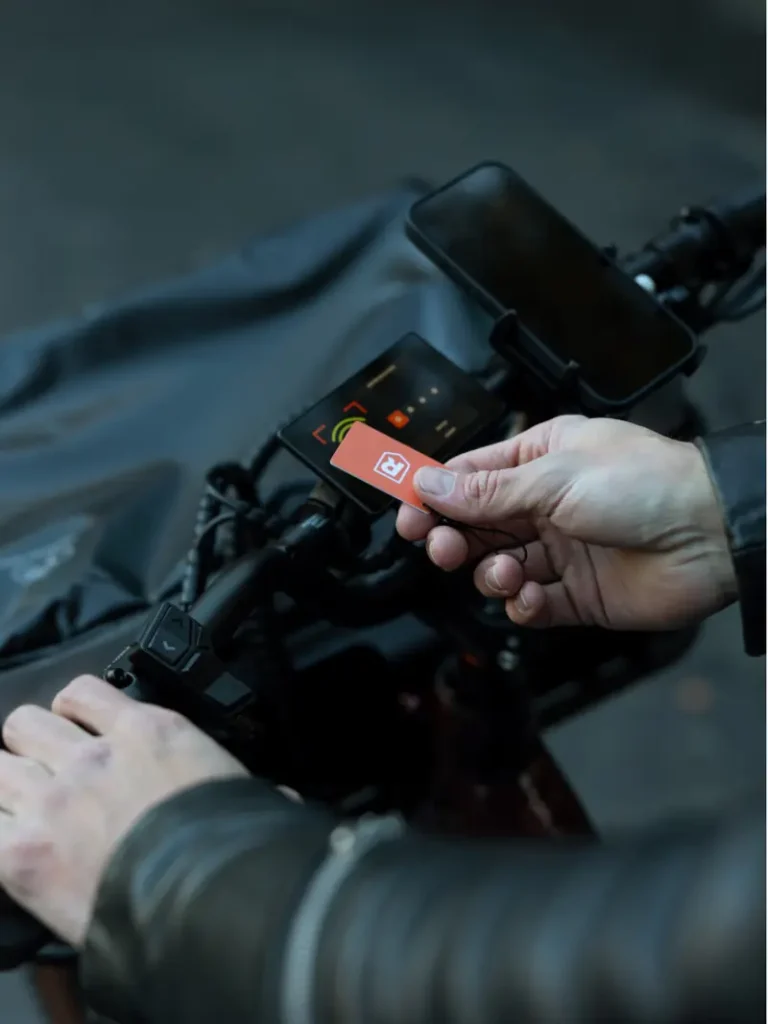 A person is holding an orange card near a reader on an electric scooter's handlebars, with a phone mounted above the display screen.