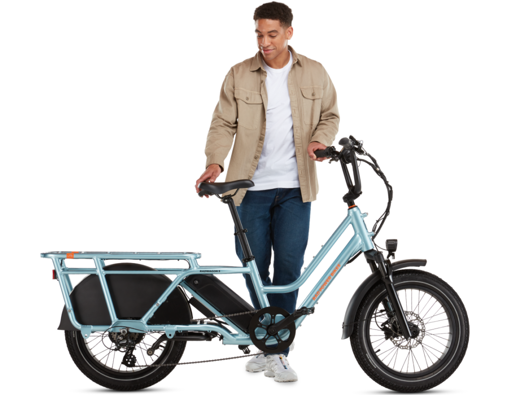 A person stands next to a blue cargo e-bike, resting one hand on the seat. The e-bike has a robust frame and large cargo area at the rear. The person wears a light brown jacket, white shirt, and jeans.