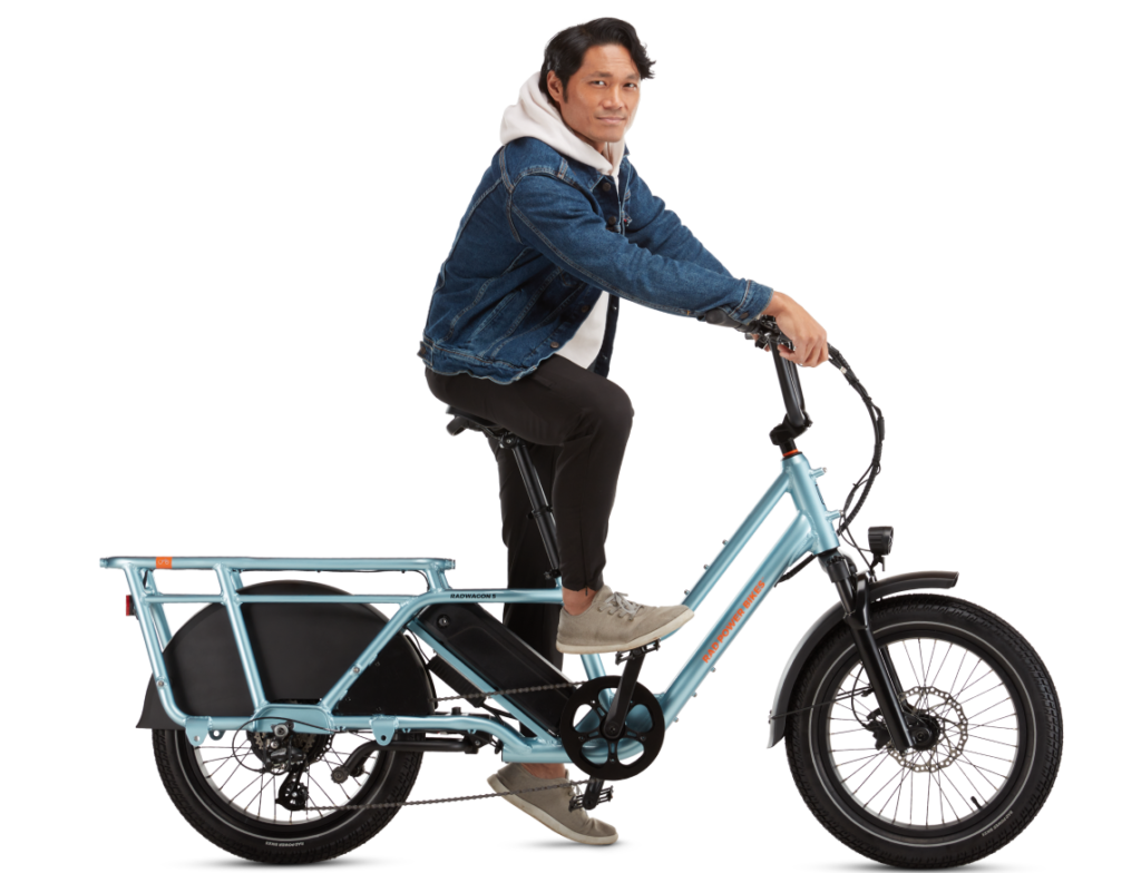 Person in a denim jacket and white hoodie sitting on a light blue electric cargo bike.