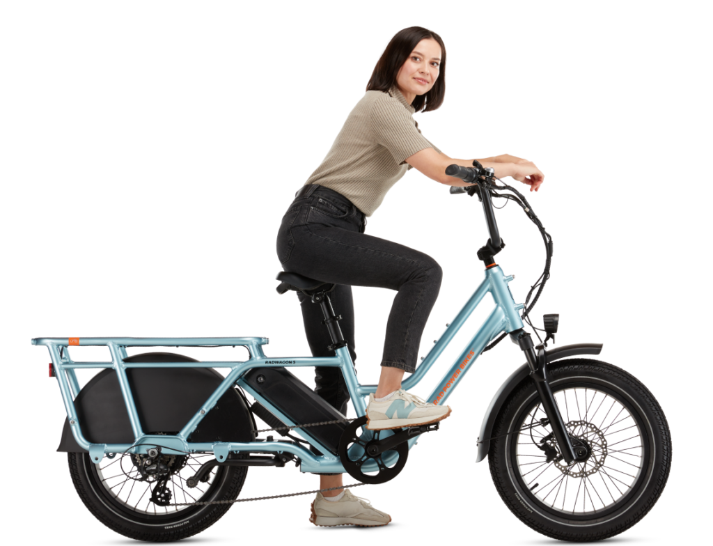 Person sitting on a blue cargo e-bike, looking ahead with hands on the handlebars. The bike features an extended rear frame and two large wheels.