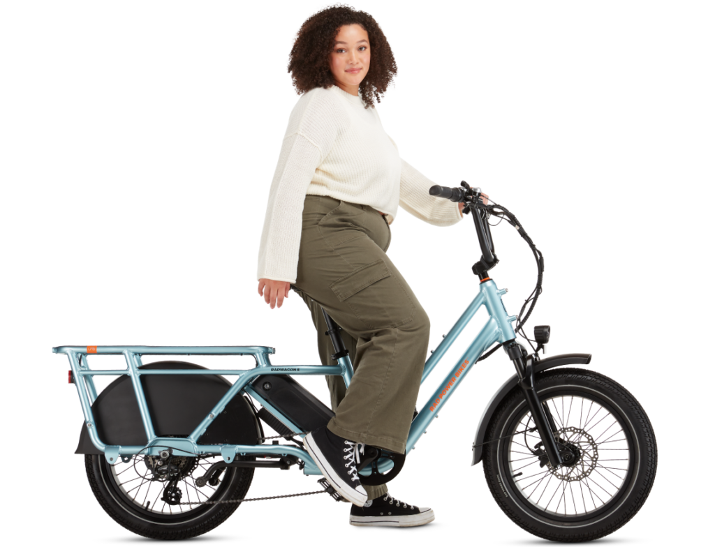 A person with curly hair, wearing a white sweater and green pants, is sitting on an electric cargo bike with a teal frame against a white background.