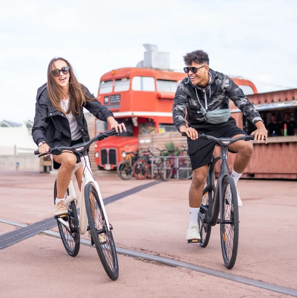 Two people, one in sunglasses and a jacket, the other in a hoodie, ride bicycles on a paved area near a red double-decker bus.