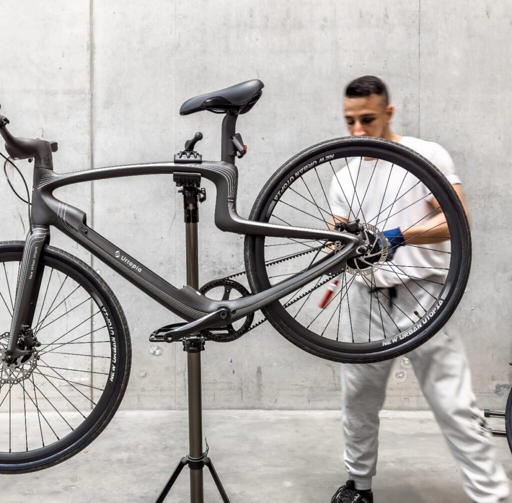 A person in a white t-shirt and grey pants works on a black bicycle mounted on a stand against a grey concrete wall.