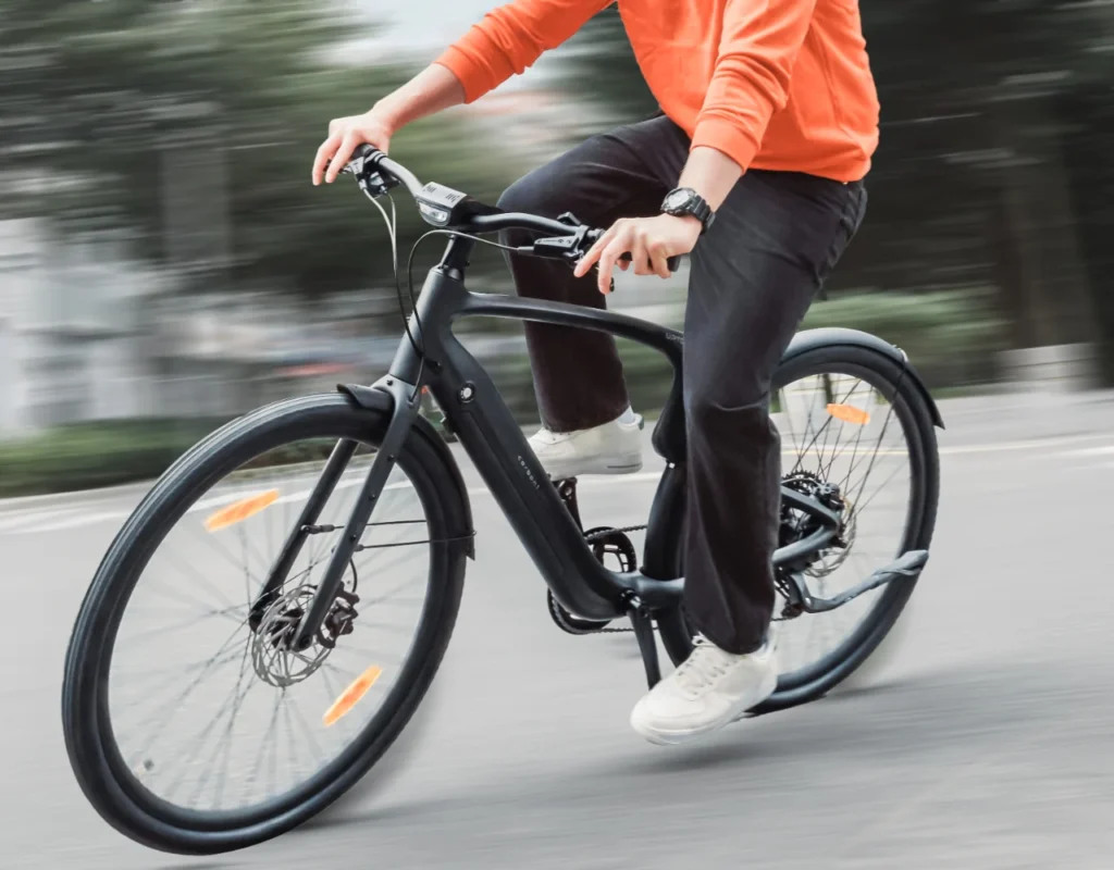 Person in an orange sweater riding a black bicycle on a smooth road, with blurred background indicating motion.