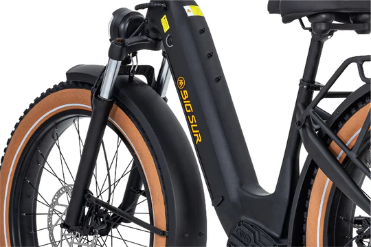 Close-up of a black electric bicycle showing the front wheel, suspension fork, and part of the frame with "Big Sur" branding. The bike features wide tires with light brown sidewalls and a rear rack.