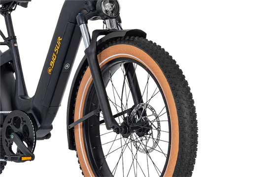 Close-up view of a bicycle's front tire and suspension fork, showcasing a thick tread and disc brake. The bike frame displays the text "GIG SUR.