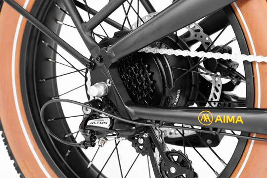 Close-up of a bicycle's rear wheel assembly, showcasing the drivetrain, disc brake, and branding with "AIMA" on the frame. The tire is tan with a textured tread pattern.