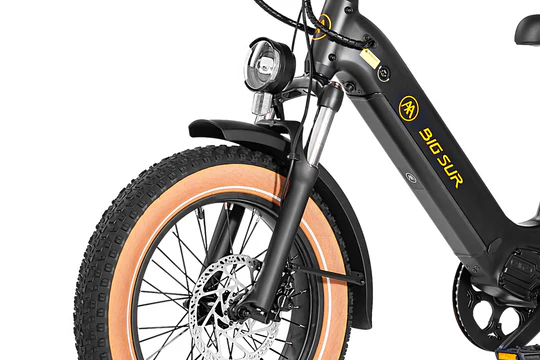 Close-up of the front section of an electric bike, showing the black frame, orange-rimmed tire, disc brake, and front light. The bike displays the brand name "Big Sur" in yellow.