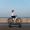 A person rides an electric bicycle along a coastal road under a clear blue sky, with "80 miles range" text displayed on the image.