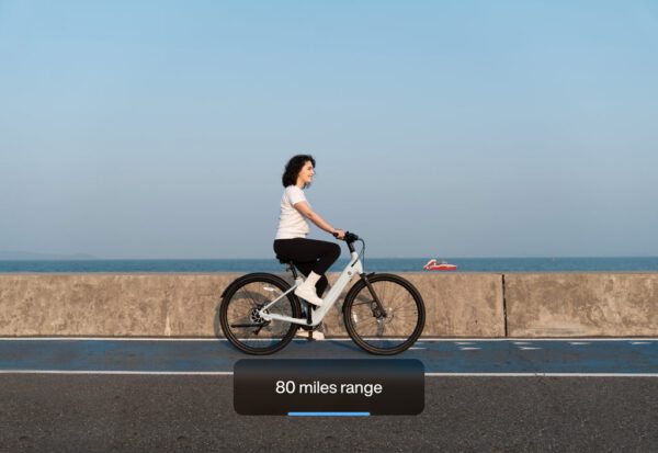 A person rides an electric bicycle along a coastal road under a clear blue sky, with "80 miles range" text displayed on the image.