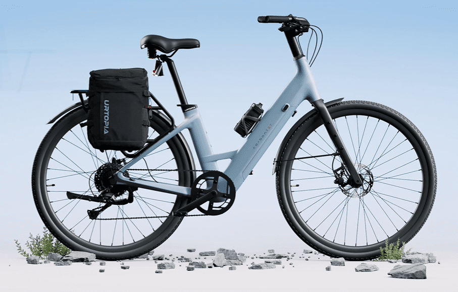 A modern, light blue electric bicycle with a black seat and a rear cargo bag, parked on a slightly rocky surface against a blue sky background.