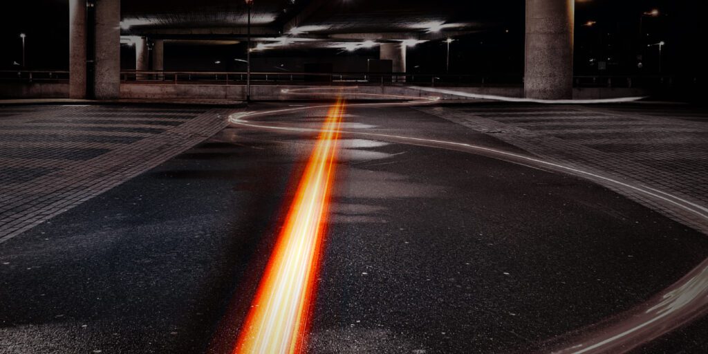 A long exposure photo shows orange light trails curving on a dark, empty road under an overpass at night.