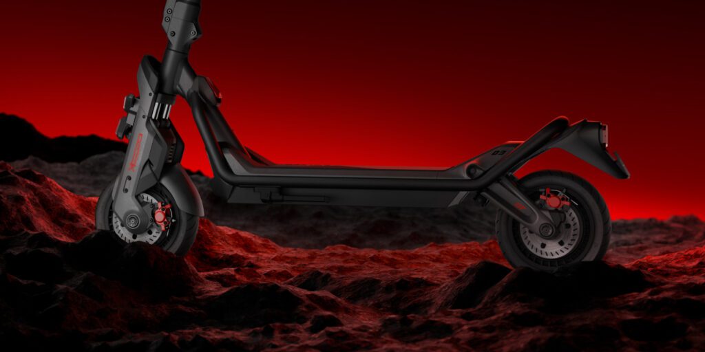 A black electric scooter is pictured in profile, positioned on rocky terrain with a red and black gradient background.