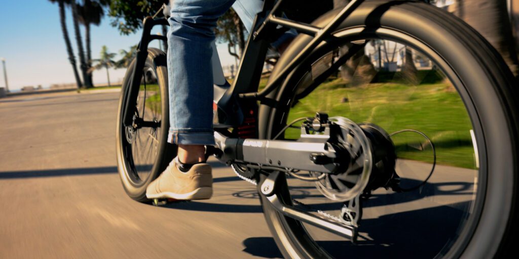 Close-up view of a person riding a bicycle on a paved path, wearing light-colored shoes and rolled-up jeans, with blurred green grass and palm trees in the background.