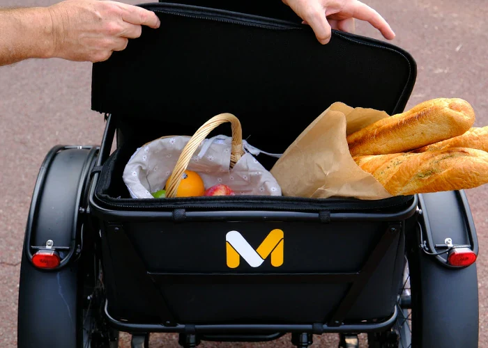 A person opens a black storage compartment on a tricycle, revealing a basket of fruit and two baguettes inside.