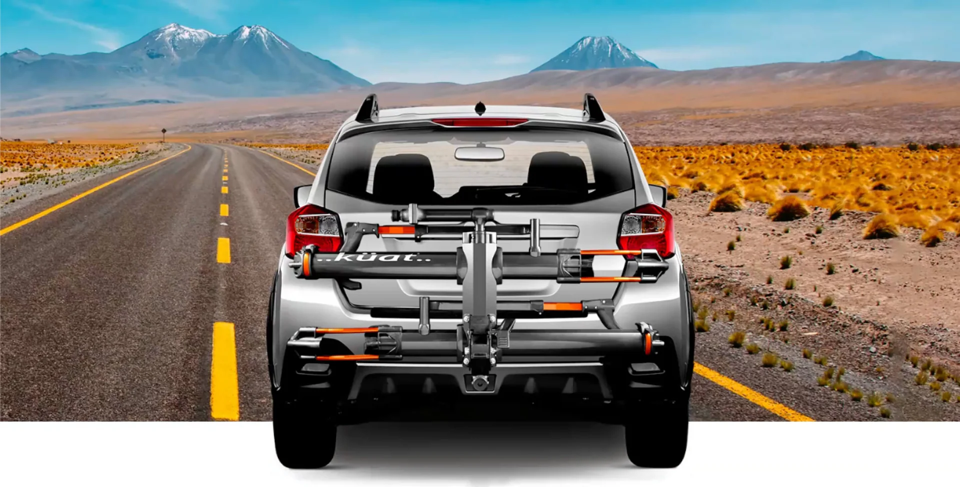A rear view of a silver SUV with a mounted bike rack on an empty desert road with mountains in the background.