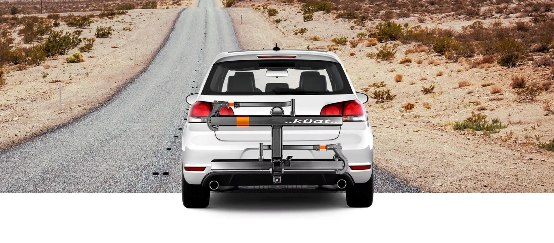 A white car with a Kuat bike rack mounted on the rear is parked on a deserted road in a dry, desert landscape.